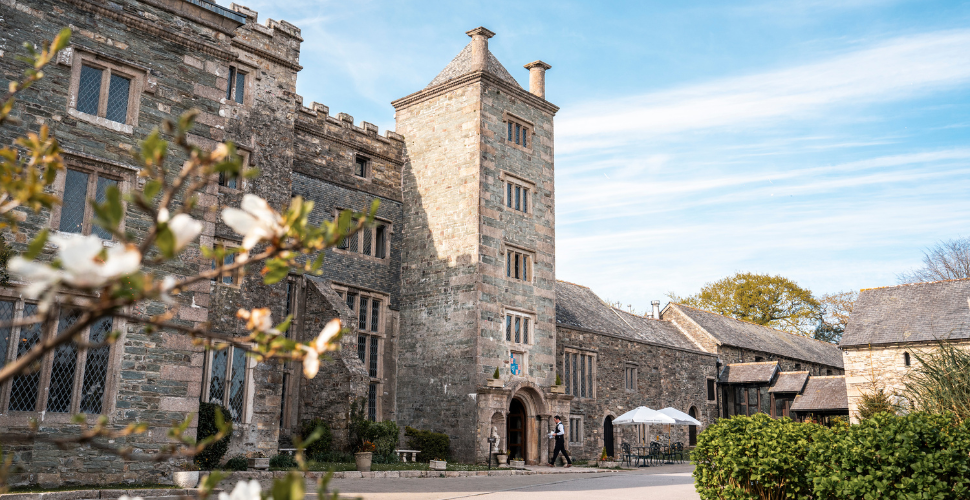 Historic stone manor house with central tower, flowering tree in the foreground, and outdoor seating under white parasols on a bright spring day.